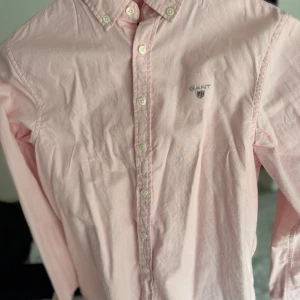 Ljusrosa skjorta från Gant Barn - Snygg ljusrosa skjorta från Gant med klassisk button down-krage och broderad logga på bröstet. Skjortan har långa ärmar och knäppning framtill med vita knappar. Perfekt för en clean och stilren look.