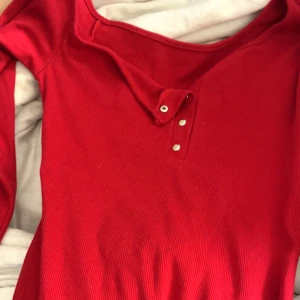 Röd ribbad långärmad klänning - Röd långärmad klänning med ribbad struktur och knappdetaljer vid halsringningen. Tröjan har en klassisk passform och är perfekt för dig som gillar en enkel men snygg stil. Materialet känns mjukt och stretchigt, vilket gör den bekväm att bära hela dagen.