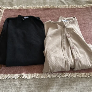 2 ribbade koftor, svart & beige, S - Två ribbade koftor i storlek S. En svart från DIVIDED och en beige från 157. Båda har lång ärm och den beiga har knappar framtill. Perfekta att slänga på sig över ett linne eller t-shirt för en chill look.