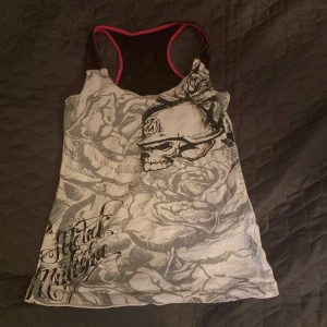 Metal Mulisha linne med dödskalle - Snyggt linne från Metal Mulisha med grått och svart dödskalletryck och rosor på framsidan. Svarta meshdetaljer och rosa kantband ger en cool kontrast. Racerback med spets upptill på ryggen. Perfekt för dig som gillar edgy streetstyle.
