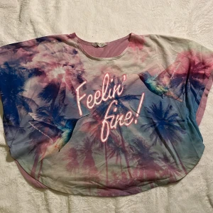 Rosa Poncho  - Säljer en loose-fit topp från H&M med coolt tryck av palmer, kolibrier och texten 'Feelin' fine!'. Framsidan har en mix av rosa, blått och lila toner, baksidan är enfärgat ljusrosa. Tunn och skön i bomull, perfekt för sommarvibbar.
