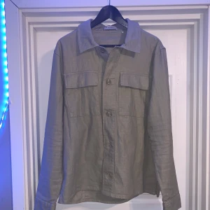Grön overshirt från Sandö - Stilren Grön overshirt från Sandö med klassisk krage och två stora bröstfickor med lock. Skjortan har knäppning framtill och är tillverkad i ett lätt och svalt tyg, perfekt för lager-på-lager. Passar dig som gillar en clean och avslappnad look.
