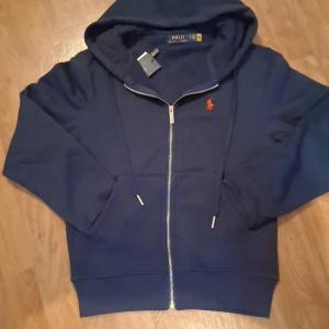 Mörkblå hoodie från Polo Ralph Lauren - Säljer en mörkblå hoodie från Polo Ralph Lauren med dragkedja och huva. Tröjan har en röd broderad logga på bröstet och är tillverkad i mjukt material. Perfekt för en avslappnad stil.