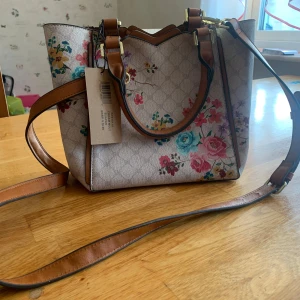 Beige axelväska med blommigt mönster - Säljer en beige axelväska från Steve Madden med färgglatt blommigt mönster och bruna detaljer. Väskan har både handtag och justerbar axelrem samt dragkedja upptill. Perfekt för dig som vill ha en snygg och praktisk accessoar.