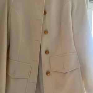 Vit kappa från Alyssa - Säljer en stilren vit kappa från Alyssa i storlek 36. Jackan har guldiga knappar framtill, två stora fickor och markerad midja baktill. Perfekt för dig som vill ha en klassisk och elegant look