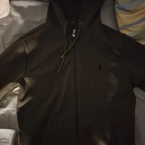 Olivgrön hoodiejacka från Polo Ralph Lauren - Säljer en olivgrön hoodiejacka från Polo Ralph Lauren med dragkedja och justerbar huva. Jackan har två fickor framtill och en broderad logga på bröstet. Perfekt för en avslappnad stil.
