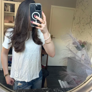 Vit t-shirt - Säljer en vit t-shirt med korta ärmar och snöre längs ena sidan. T-shirten har en avslappnad passform och är tillverkad i ett lätt bomullsmaterial. Perfekt till jeans för en enkel och stilren look. Den har tryck med vit text på sig.