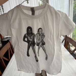 Vit t-shirt med svart tryck XS - Vit t-shirt i bomull med coolt svart tryck framtill som föreställer två personer i sittande pose. Klassisk rund halsringning och korta ärmar. Perfekt för dig som gillar streetstyle och grafiska motiv.