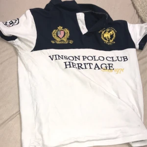 Vit och marinblå pikétröja från Vinson Polo Club - Säljer en stilren pikétröja från Vinson Polo Club i vitt och marinblått med broderade detaljer och loggor i gult och rött. Tröjan har krage, korta ärmar och tryckt text på bröstet. Perfekt för en sportig look.