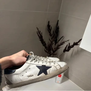  Golden Goose sneakers  - Säljer ett par vita sneakers från Golden Goose med en svart stjärna på sidan och grå snörning. Skorna har rund tå och platt sula, samt detaljer i skinn. Perfekta för en avslappnad stil.
