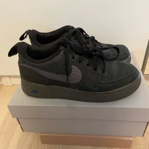 Nike Air Force 1 GS svart/grå - Säljer ett par Nike Air Force 1 GS i svart med grå detaljer och blå mini-swoosh. Klassisk low-top siluett med snörning och rund tå. Ovandel i skinn och syntet, robust sula och broderad Nike Air-logga på hälen. Perfekt för dig som gillar streetstyle. Finns smått slitage på insidan av swooshen men ingen som märks.