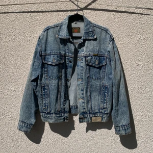 Blå jeansjacka från Wrangler - Klassisk blå jeansjacka från Wrangler med två bröstfickor med lock och knappar samt sidofickor. Jackan är lätt bomber i passform och tydliga sömmar i kontrastfärg. 
