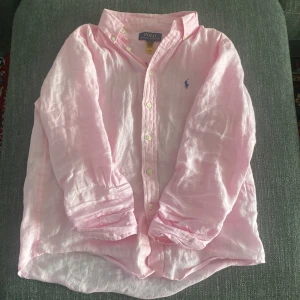 Rosa linneskjorta från Polo Ralph Lauren - Ljusrosa skjorta från Polo Ralph Lauren i luftig linnekvalitet. Klassisk button-down krage, långa ärmar och broderad blå logga på bröstet. Perfekt för en chill och stilren look, lätt att matcha med jeans eller shorts. Men behöver strykas så är den som ny.