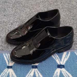 Svarta lackloafers från Ellos - Snygga svarta loafers i lack från Ellos med elastisk detalj över vristen. Skorna har rund tå, platt sula med grovt mönster undertill och en stilren look som passar till många outfits. Perfekta för dig som gillar klassisk stil med en modern twist. Skor är nästan nya, används bara en gång. 