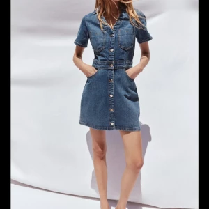 Jeansklänning med knappar - Snygg jeansklänning i blå denim med korta ärmar, knappar framtill och fickor både fram och bak. Klänningen har en klassisk skjortkrage och markerad midja för en avslappnad men trendig look.