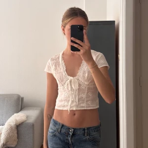 Vit spetsig croppad blus - Söt vit croppad blus med spetsdetaljer och broderat blommönster. Djup v-ringning och knyt framtill, kortärmad och lätt transparent. Perfekt till jeans eller kjol för en somrig look.💐
