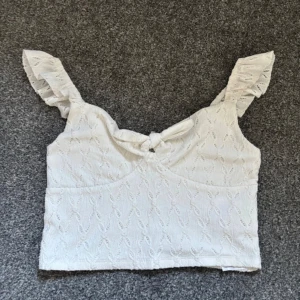 Croppas topp med volangärmar - Säljer en söt vit croppad topp från FB Sister med volangärmar och knytdetalj framtill. Toppen har ett diskret broderat mönster och är perfekt för varma dagar. Supersnygg att matcha med jeans eller kjol!