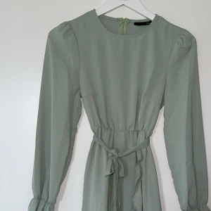 Ljusgrön klänning med volang och knytband - Säljer en ljusgrön klänning med långa ärmar, markerad midja och knytband. Klänningen har volangdetaljer längs nederkanten och är tillverkad i ett lätt, flowy material. Perfekt för vår och sommar!