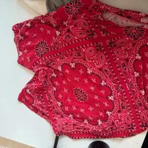 Röd mönstrad klänning från Bik Bok - Säljer en röd klänning från Bik Bok med bandana-inspirerat paisleymönster i svart och vitt. Klänningen har korta ärmar och resår i midjan för en bekväm passform. Perfekt för dig som gillar färgstarka och mönstrade plagg!