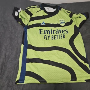 Arsenal bortatröja Xhaka 34 - Säljer en Arsenal bortatröja från Adidas med Xhaka 34 på ryggen. Tröjan är ljusgul med svarta och blå detaljer samt randigt mönster. Tryck med Emirates Fly Better och Visit Rwanda på ärmen. Premier League-märke på ena ärmen.