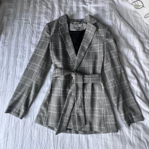 Klassisk rutig kavaj med midjeskärp - Stilren kavaj från H&M i grått och svart rutmönster. Markerad midja med avtagbart tygbälte för en figursydd look. Passar perfekt till både jobb och fest. Storlek: S Passform: Normal Fodrad på insidan och med snygg krage. Perfekt för dig som vill ha en klassisk men trendig stil.