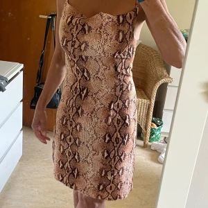 Ormmönstrad kortklänning från Boohoo - Säljer en snygg kortklänning från Boohoo med ormmönster i bruna och beige toner. Klänningen har smala axelband och figurnära passform, perfekt för dig som vill sticka ut. Materialet är mjukt och följsamt.