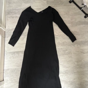 Svart ribbad långklänning - Svart långklänning med ribbad struktur och v-ringning. Klänningen har långa ärmar och en smickrande passform som följer kroppen. Perfekt för dig som gillar en stilren och enkel look.