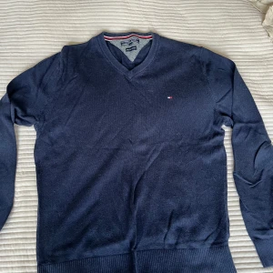 Mörkblå v-ringad tröja Tommy Hilfiger - Mörkblå v-ringad tröja från Tommy Hilfiger i en mjuk mix av pima bomull och kashmir. Klassisk design med diskret logga på bröstet och ribbade muddar. Perfekt för dig som gillar stilrena och bekväma plagg.