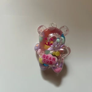 Pastellfärgat björn-nyckelring BEAR CUB - Supergulligt nyckelring med en genomskinlig björnberlock fylld med pastellfärgade pärlor och texten 'BEAR CUB' i rosa. Berlocken har inslag av lila, rosa, blått, gult och grönt och är perfekt för dig som gillar söta accessoarer med lekfull stil.