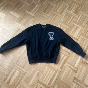 Svart sweatshirt från Ami Paris - Svart sweatshirt från Ami Paris med ikonisk hjärtformad A-logga på bröstet. Tröjan har rund hals, ribbade muddar och är tillverkad i mjuk bomull. Perfekt för dig som gillar stilrena och trendiga plagg med en twist.