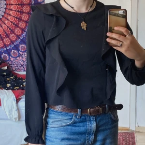 Svart blus med volanger från KappAhl, M - Svart blus från KappAhl med långa ärmar och snygga volangdetaljer längs framsidan och i slutet av ärmarna. Den har ett lätt material som känns skönt mot huden. Använt kanske två gånger, nyskick.