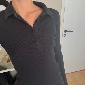 Svart ribbad polotopp med krage - Säljer en svart ribbad polotopp med krage och knappar framtill. Tröjan har lång ärm och är figurnära i modellen. Perfekt till både jeans och kjol för en stilren look.