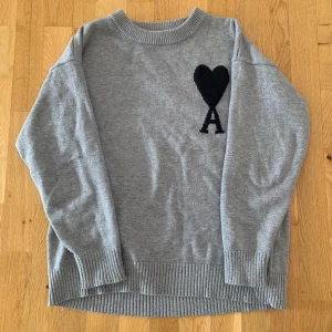 Grå stickad Ami Paris - Säljer en grå stickad tröja från Ami Paris med svart hjärta och bokstaven A på bröstet. Tröjan har rund halsringning, långa ärmar och ribbade muddar vid ärmslut och nederkant. Perfekt för dig som gillar stilrena plagg med en twist.
