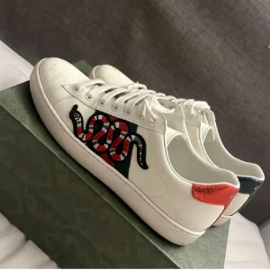 Gucci sneakers med bi - Säljer ett par vita Gucci sneakers med klassiska gröna och röda ränder på sidan samt en broderad bi. Skorna har snörning och detaljer i rött och grönt på hälen. Snygg och unik design som sticker ut. Skorna har ett bi och inte en orm!