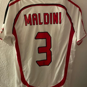 AC Milan Maldini Champions League tröja - Säljer en ikonisk AC Milan bortatröja från Champions League-finalen 2007 med Maldini #3 på ryggen. Tröjan är vit med röda och svarta detaljer, Adidas-logga, klubbmärke och Bwin som sponsor. Tillverkad i lätt och ventilerande material, perfekt för fotbollsfans.