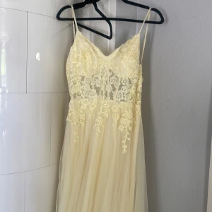 Ljusgul långklänning från JJ's House - Superfin ljusgul långklänning från JJ's House med tunna axelband och vacker spets upptill. Klänningen har blommiga broderier på livet, v-ringning och ett flowy, tunt tyg i kjolen. Perfekt för dig som vill sticka ut med en romantisk look.