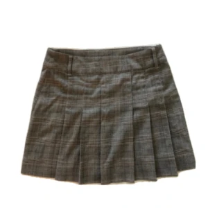 Grå rutig kortkjol med dragkedjor - Snygg grå rutig kortkjol med veckad design och bälteshällor. Kjolen har två dekorativa dragkedjor baktill och klassiskt rutmönster. Perfekt för en trendig och stilren look.