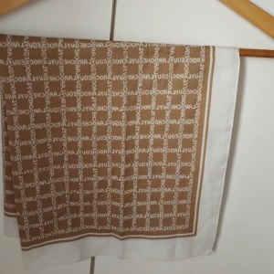 Beige och vit scarf från Guy Laroche - Säljer en snygg beige och vit scarf från Guy Laroche Paris. Scarfen har ett grafiskt mönster med varumärkets namn tryckt över hela tyget och en vit kant runtom. Perfekt accessoar för att lyfta din outfit.