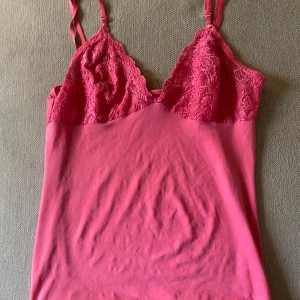 Vintage rosa topp med spets  - Supersöt rosa linnetopp med tunna justerbara axelband och v-ringad spets upptill. Toppen har en slät nederdel i mjukt tyg och detaljerad spets över bysten som ger en romantisk vibe. Perfekt att styla med jeans eller kjol för en trendig look.