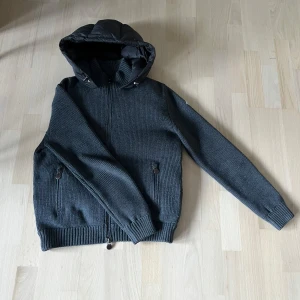 Moncler cardigan  - Snygg grå dunjacka från Moncler med stickade ärmar och ribbade muddar. Jackan har svart quiltad rygg och huva, dragkedja framtill och två fickor med dragkedja. Perfekt för kalla dagar när du vill ha både stil och värme. Skriv för fler bilder!