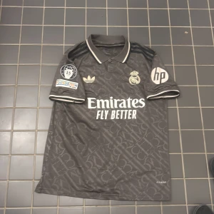 Real Madrid svart fotbollströja från Adidas - Säljer en grå Real Madrid fotbollströja från Adidas med vita detaljer och tryck. Tröjan har korta ärmar, pikékrage och klubbmärke på bröstet. Sponsorer som Emirates och HP syns tydligt. Perfekt för fans!