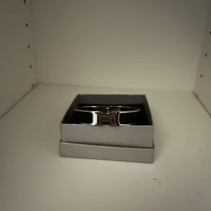 Silverfärgat armband med H-detalj - Snyggt armband i silverfärg med en tydlig H-detalj framtill. Perfekt accessoar för att lyfta din outfit med en stilren och modern touch. Kommer i en elegant ask.