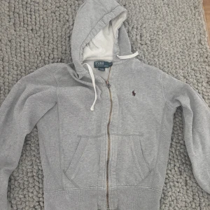 Grå hoodie från Polo Ralph Lauren - Säljer en klassisk grå hoodie från Polo Ralph Lauren med dragkedja och huva. Tröjan har fickor framtill, vit dragsko och den ikoniska lilla broderade loggan på bröstet. Perfekt för en avslappnad stil.