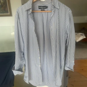 Randig skjorta från Ralph Lauren - Klassisk skjorta från Ralph Lauren i blåvitt randigt mönster. Skjortan är i 100% bomull, har långa ärmar och knappar framtill. Broderad logga med polospelare på bröstet. Perfekt för en clean och stilren look.