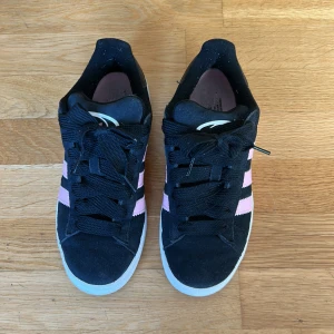 Adidas Campus sneakers svart med rosa ränder - Säljer ett par Adidas Campus sneakers i svart mocka med klassiska rosa ränder på sidorna och vit sula.                                                                        Bra skick på utsidan, inga synliga fläckar vad jag märkt. Använda ett tiotal gånger, insidan är sliten i sulorna och lite luddig i härlarna men kan fräschas upp.