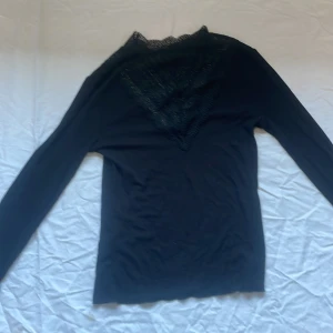 Svart långärmad topp med spetsdetaljer - Säljer en svart långärmad topp med vacker spets runt halsringningen och ärmsluten. Toppen har en elegant v-ringad spets framtill och är tillverkad i ett mjukt, stretchigt material. Perfekt för dig som gillar detaljer och vill ha något extra till din outfit.