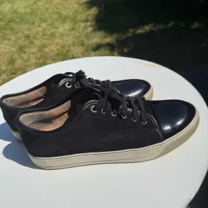  Lanvin  - Säljer ett par svarta sneakers från Lanvin med snygg lackad tå och vit sula. Skorna har snörning och en klassisk, stilren design som passar till mycket. Märket syns på innersulan och yttersulan. Perfekta för dig som gillar exklusiva sneakers. Box dusbag och snoskören medföljer 