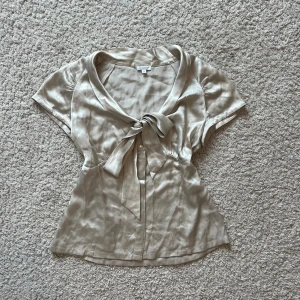 Fest topp/blus - Snygg champagnefärgad blus i glansigt satinliknande material. Blusen har korta ärmar, v-ringning och en stor knytrosett framtill. Knappar längs hela framsidan och figurnära passform. 💖Behöver bara strykas lite men annars så fin!