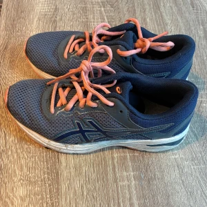 Blå Asics sneakers med rosa snören - Säljer ett par blå Asics sneakers med mesh-ovandel och rosa snören. Skorna har en vit och grå sula med dämpning och klassisk Asics-logga på sidan. Perfekta för träning eller en aktiv vardag. Snygg sportig look och riktigt bekväma på foten. Storlek 36 och är 22.5 cm.