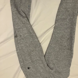 Grå mjukisbyxor från Champion - Gråa mjukisbyxor från Champion med klassisk logga på benet. Byxorna är i mjukt bomullsmaterial och har en avslappnad passform. Perfekta för chill dagar eller när du vill vara bekväm. Elastisk midja och muddar vid benslut.